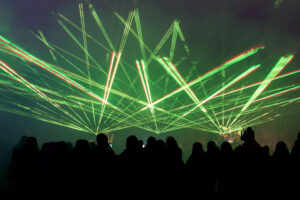 Silhouettes of people watching a vibrant green laser display event inside a dark venue.
